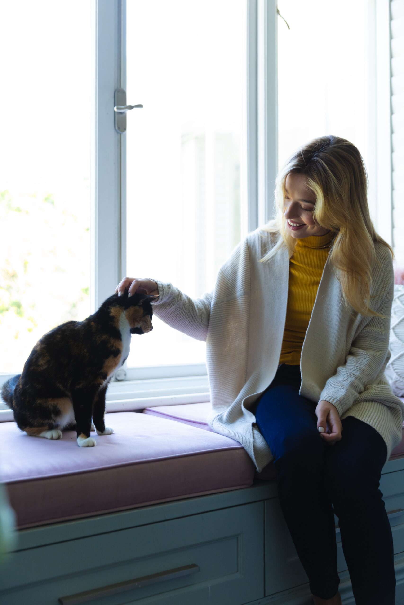 Happy caucasian woman sitting on windowsill and stroking cat in sunny room at home. Relaxation, wellbeing, cat, pet, emotions,calm, free time and domestic life, unaltered.